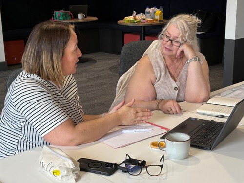 Two women sat by a laptop talking.