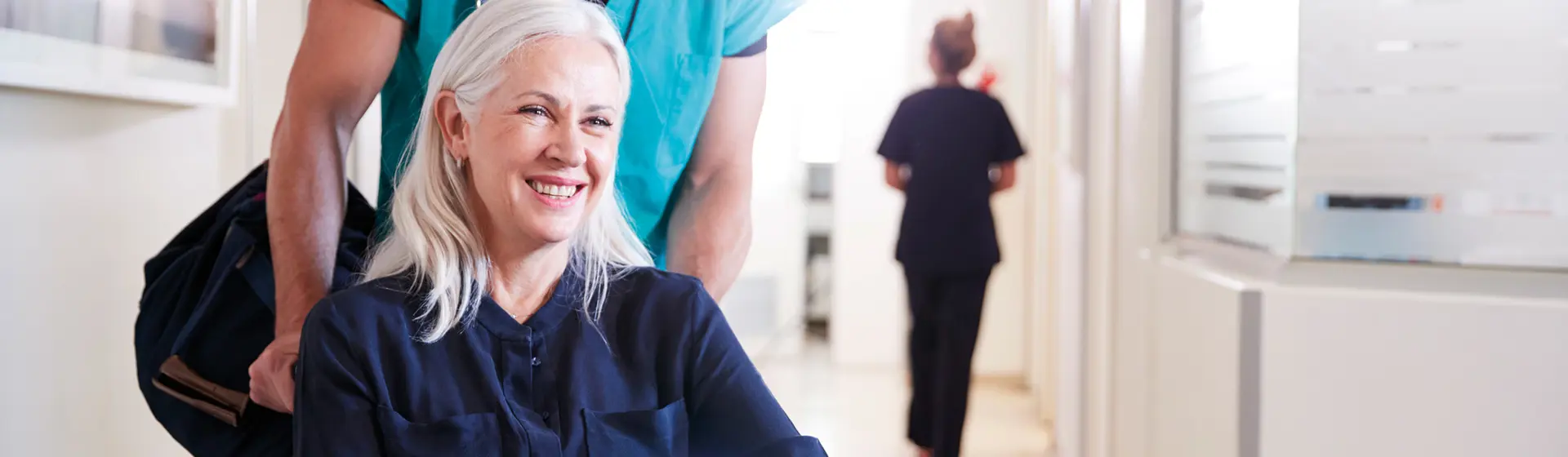 An elderly lady with grey hair being pushed in a wheelchair by a smiling man