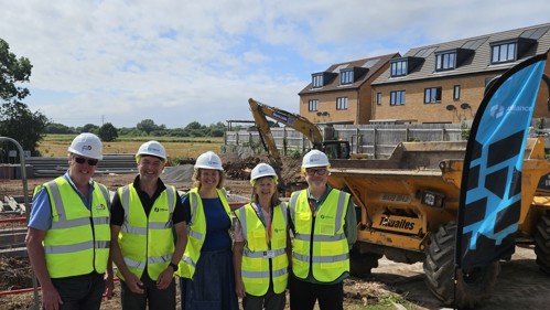 Five people at a constructin site wearing hi-viz vests. A dumper truck and digger are in the background.