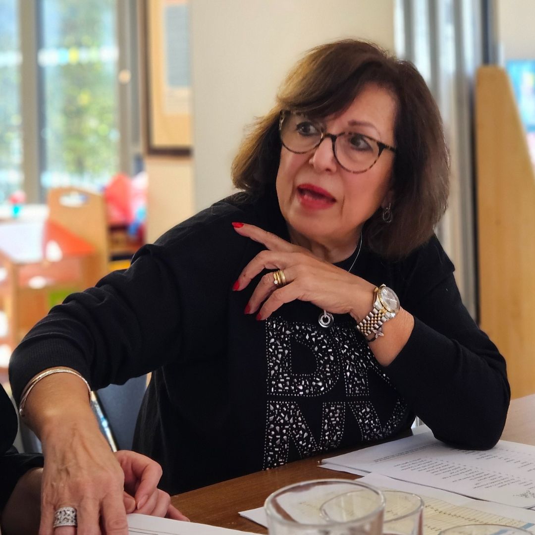 A middle-aged women talking to someone off screen sat at a desk.