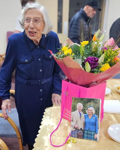 A lady with silver hair wearing glasses and a blue jump suit stood next to a vase of flowers.