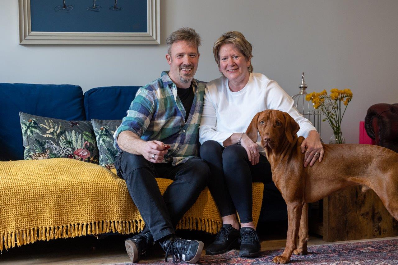 A couple sit on a sofa with a dog next to them