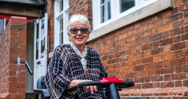 Women On A Mobility Scooter Outside, Sat In Front Of A Red Brick Wall