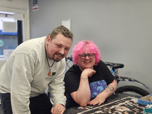 Two people looking at the camera, one of the people featured has bright pink hair.