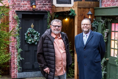 Two men standing together against a Victorian-themed Christmas street scene.